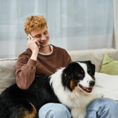 A young man with red hair chats on the phone while relaxing with his dog on the couch.